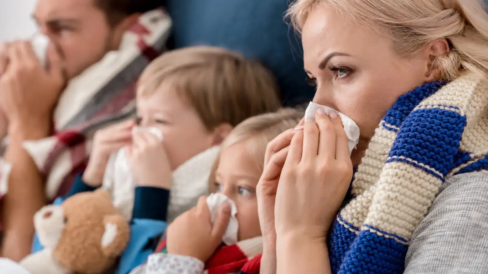 A family of four wrapped in blankets on a sofa, all blowing their noses into tissues
