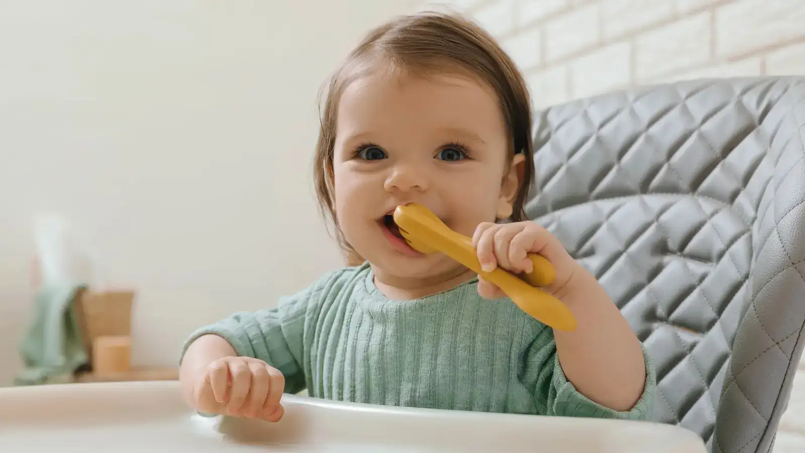 A baby in a highchair chewing on a yellow plastic spoon