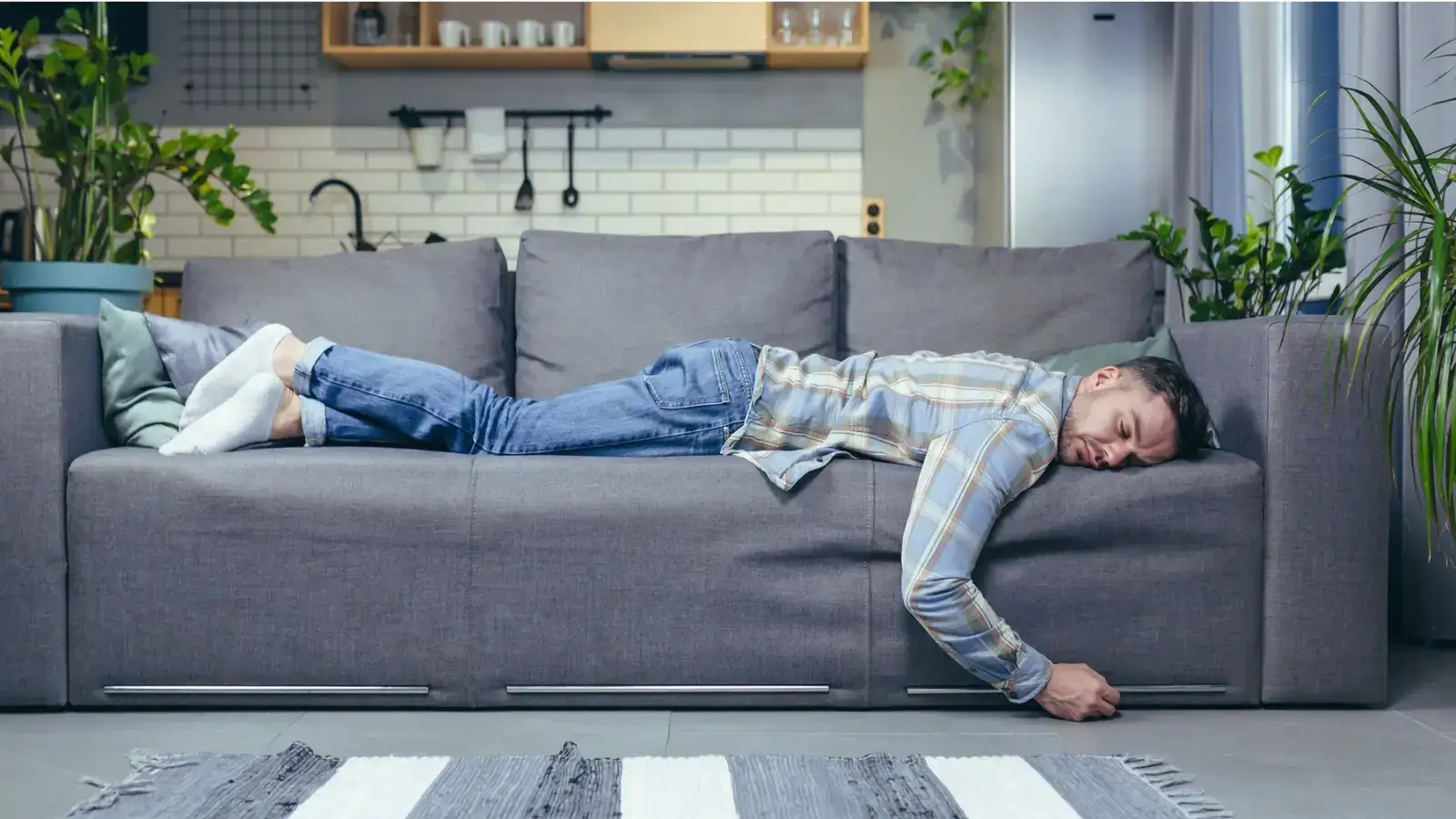 A man lying face down across a grey sofa in a living room, one arm hanging to the floor