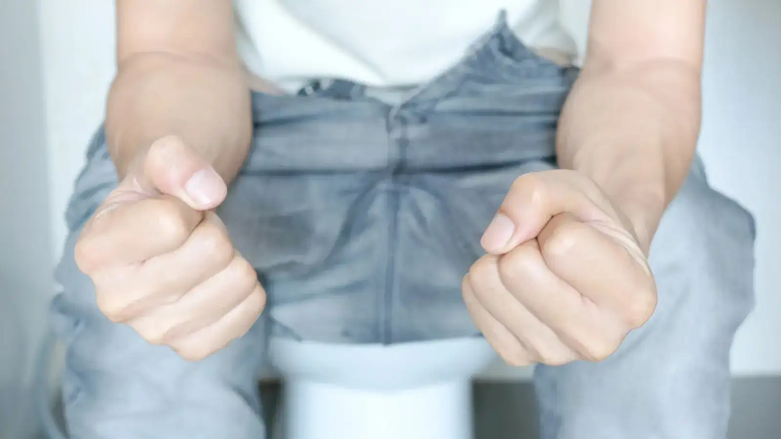 A person sitting on a toilet in jeans with both fists clenched on their knees