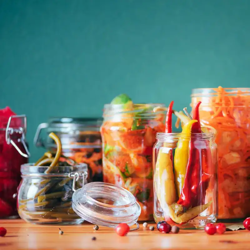 Jars of fermented chillies, carrots and cucumbers on a wooden table against a teal background