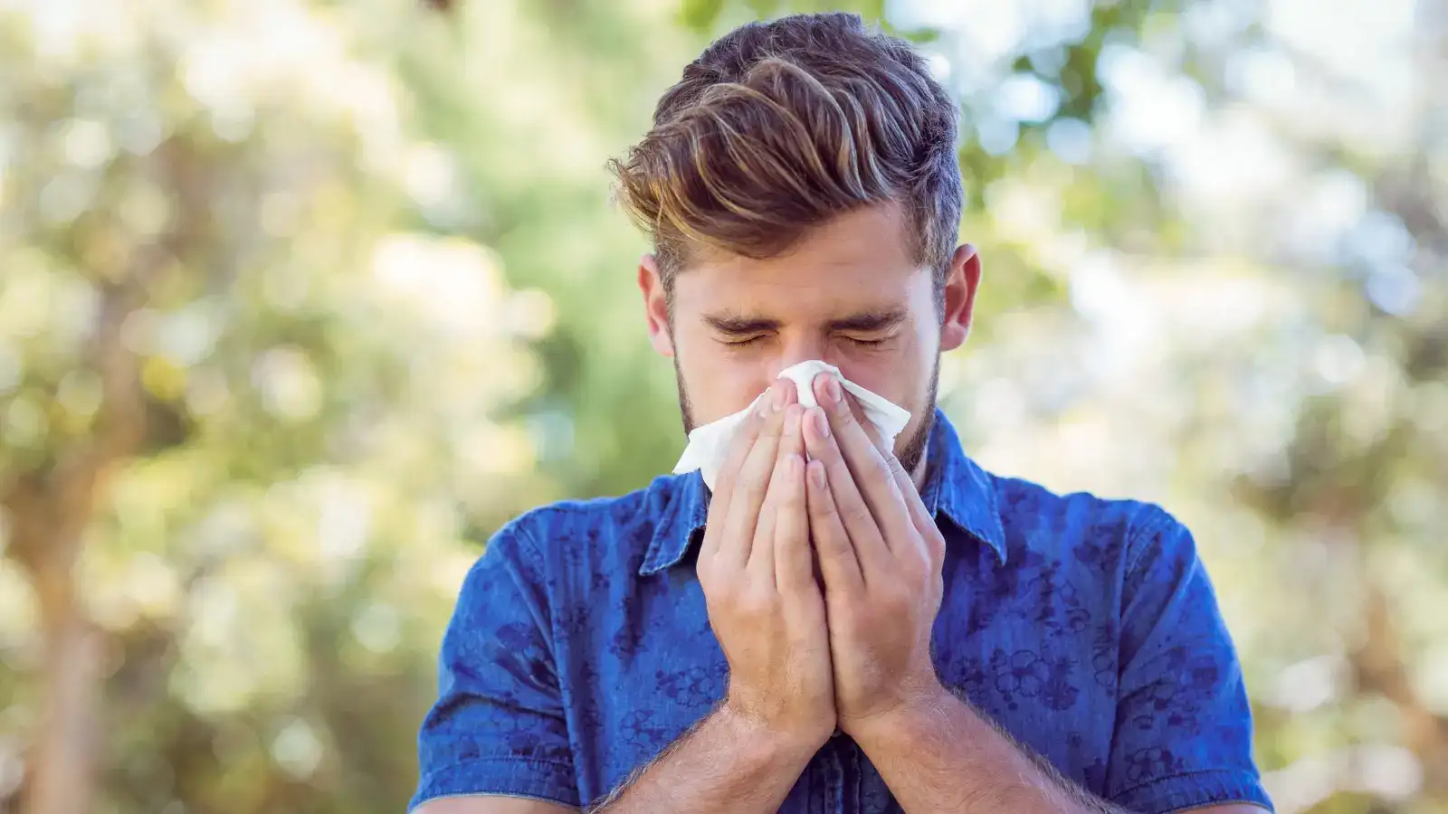 A man in a blue shirt blowing his nose into a tissue outdoors among greenery