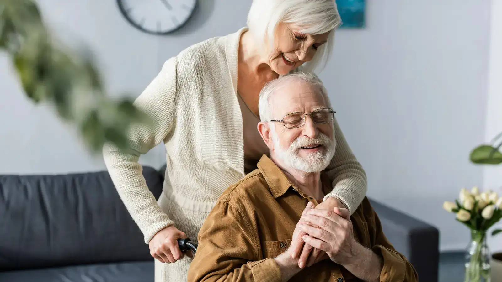 An older man sitting with his hands clasped while a woman rests a hand on his shoulder