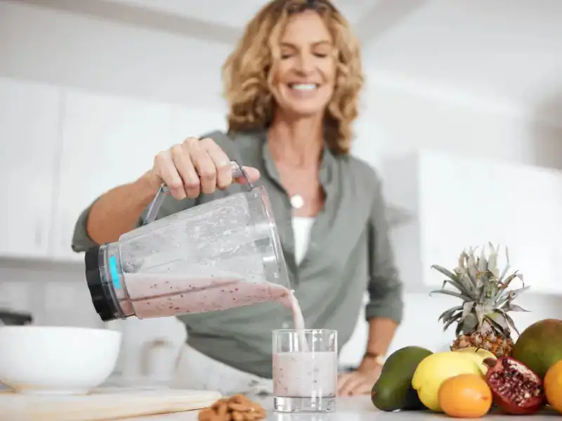 A woman pouring a smoothie from a blender jug into a glass beside a bowl of fruit