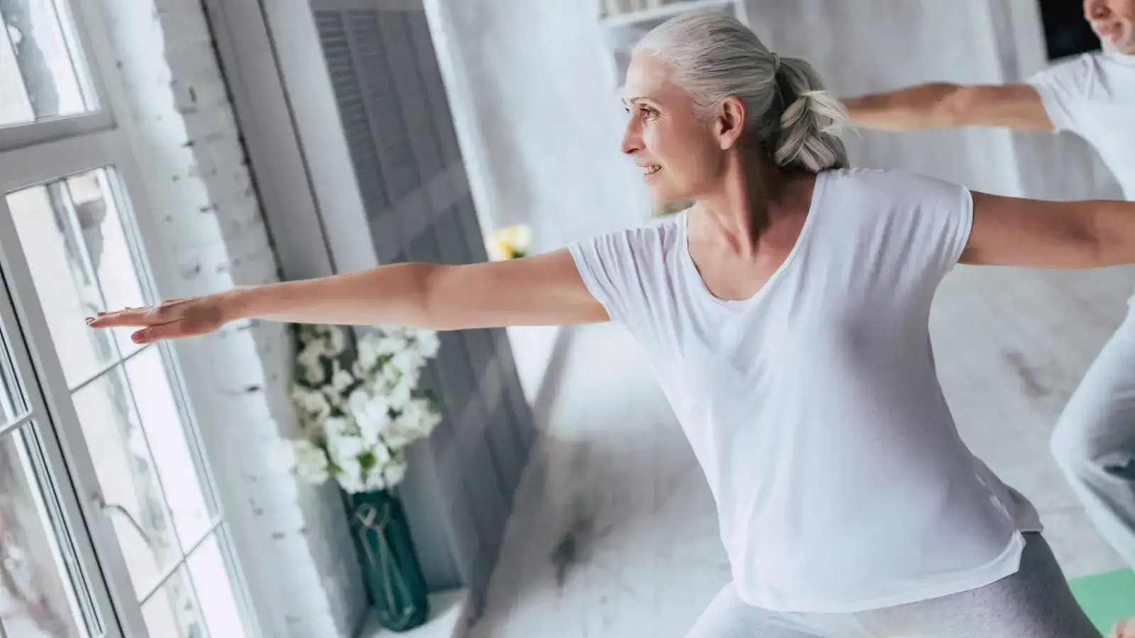 An older woman in a white t-shirt stretching with both arms out beside a window