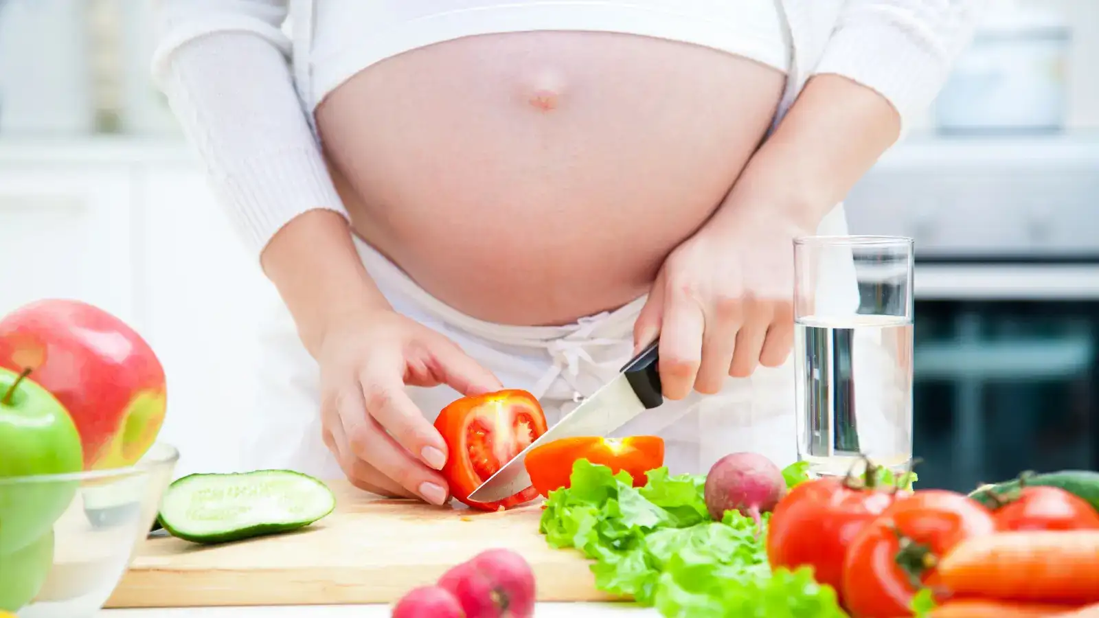 A pregnant woman slicing peppers at a kitchen bench beside fruit and vegetables